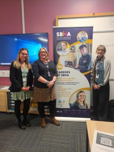 Three women standing in front of a roll-up banner for the Scottish Borders Housing Association (SBHA). The banner features the SBHA logo, photos of staff members, and text promoting careers at SBHA, including opportunities in trades, housing and tenancy management, customer services, technical services, and support services. The women are smiling, dressed in professional attire, and standing in a classroom or office setting with a whiteboard and a computer monitor in the background.