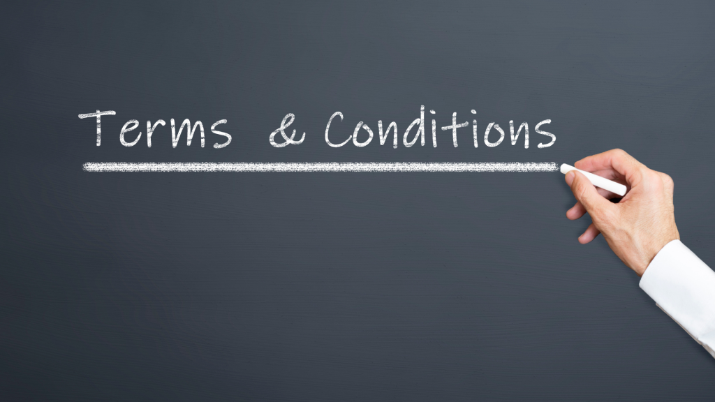 The image shows a dark grey chalkboard with the words "Terms & Conditions" written in white chalk. A hand, dressed in a white shirt sleeve, is seen holding a piece of chalk and drawing an underline beneath the text.