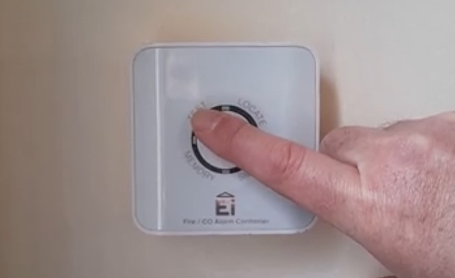 A close-up image of a person's hand pressing a button on a wall-mounted Fire/CO Alarm Controller. The device has a square design with rounded edges and features a central circular button with labels such as "TEST," "LOCATE," and "MEMORY."